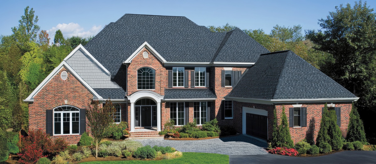 Modern residential architecture featuring a stylish black shingled roof on a brick home surrounded by lush green trees and a paved driveway.